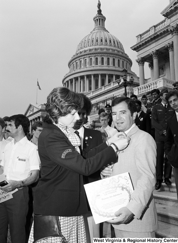 Congressman Rahall stands on the steps of the Capitol Building holding a diploma presented by L'AERO CLUB DE FRANCE, while a woman wearing a "FRANCE" insignia pins something to his lapel.