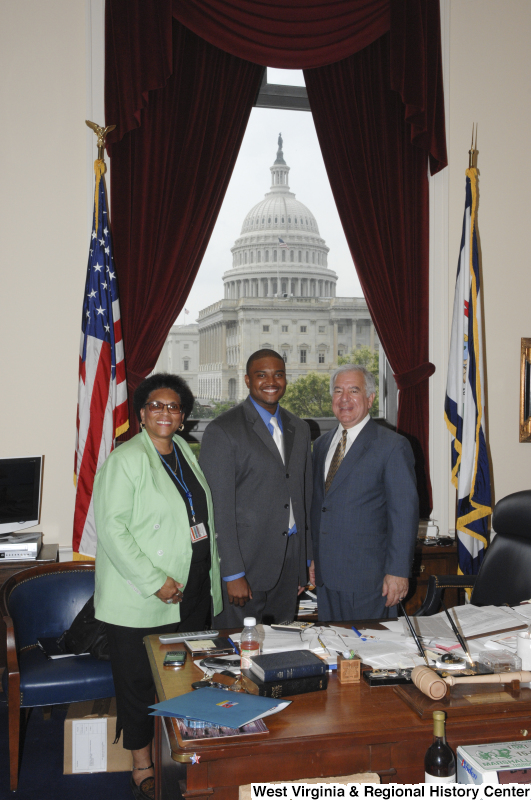 Congressman Rahall stands in his Washington office with a man wearing a grey suit, blue shirt, and light tie, and a woman wearing a light green sport coat.