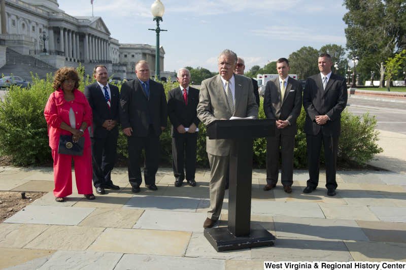 A man stands at a podium with Congressman Rahall and other people during an outdoor press conference near the Capitol Building.