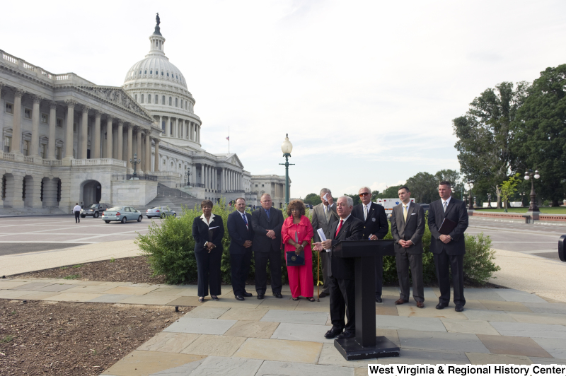 Congressman Rahall stands at a podium with eight other people during an outdoor press conference near the Capitol Building.