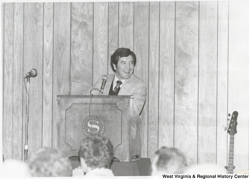 Photograph of Congressman Nick Rahall II standing at a podium speaking to a crowd. The podium has an "S' with a laurel wreath around it.