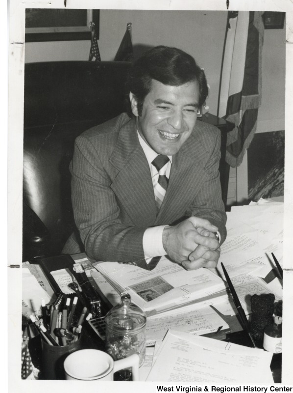 Photograph of Congressman Nick Rahall II sitting at his desk laughing. His desk is covered in papers and notes.