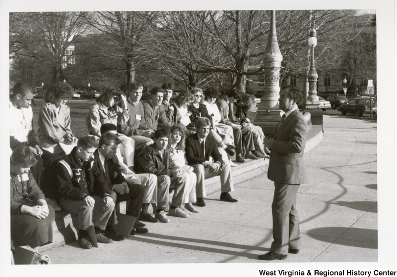Congressman Nick Rahall II speaking to a group of high school students sitting in front of him. Rahall is standing on a sidewalk in front of the U.S. Capitol Complex.