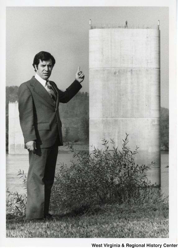 Congressman Nick Rahall II standing by a river pointing at two cement bridge supports  in the water, possibly in Huntington, West Virginia.
