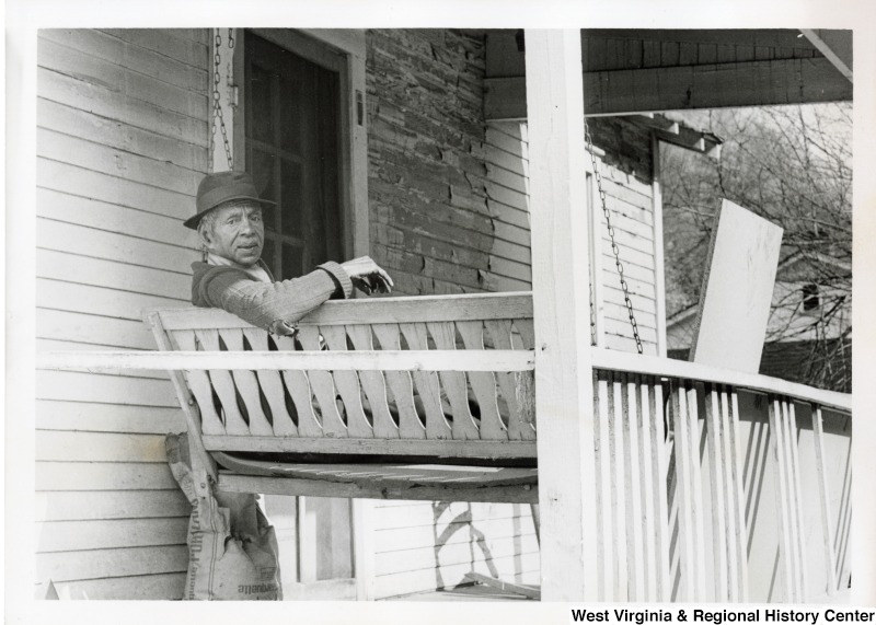 An unidentified man sitting on a porch swing in Mingo county. He is looking over his shoulder at the photographer. Some flood damage to the house can be seen behind the man.