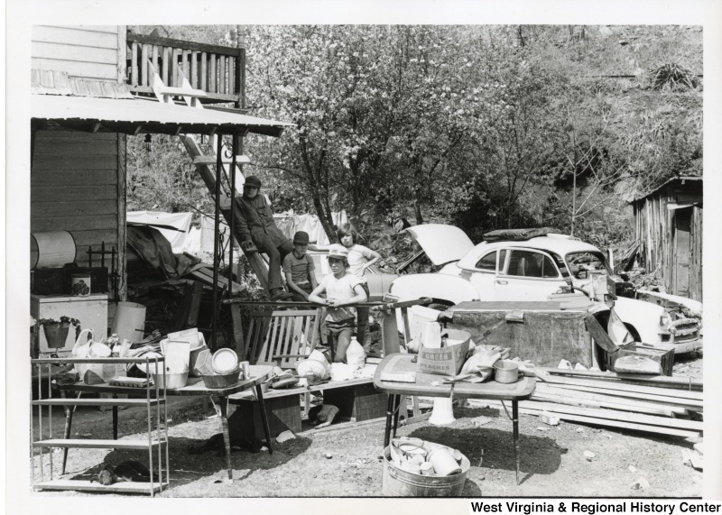 Four unidentified boys are standing by a ladder leading to the second story of a flood damaged house in Mingo county. They are surrounded by household items that were damaged or salvaged from the flood. A broken down car is behind them.