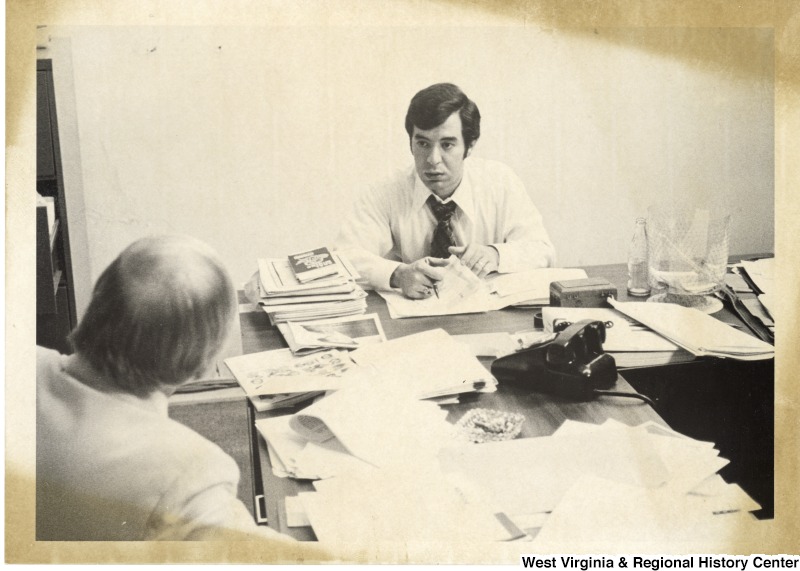 Congressman Nick Rahall II seated at a desk talking to an unidentified man.