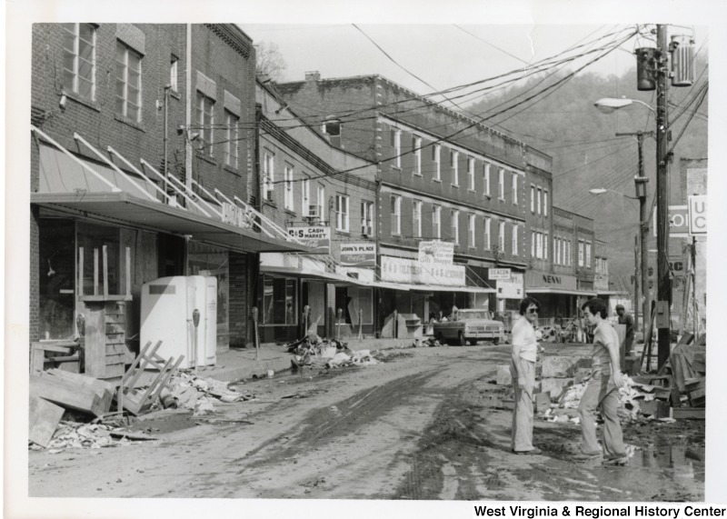 A flood damage street in Mingo county. There are two unidentified men in the right corner of photograph. The street is littered with debris and damaged goods from stores.