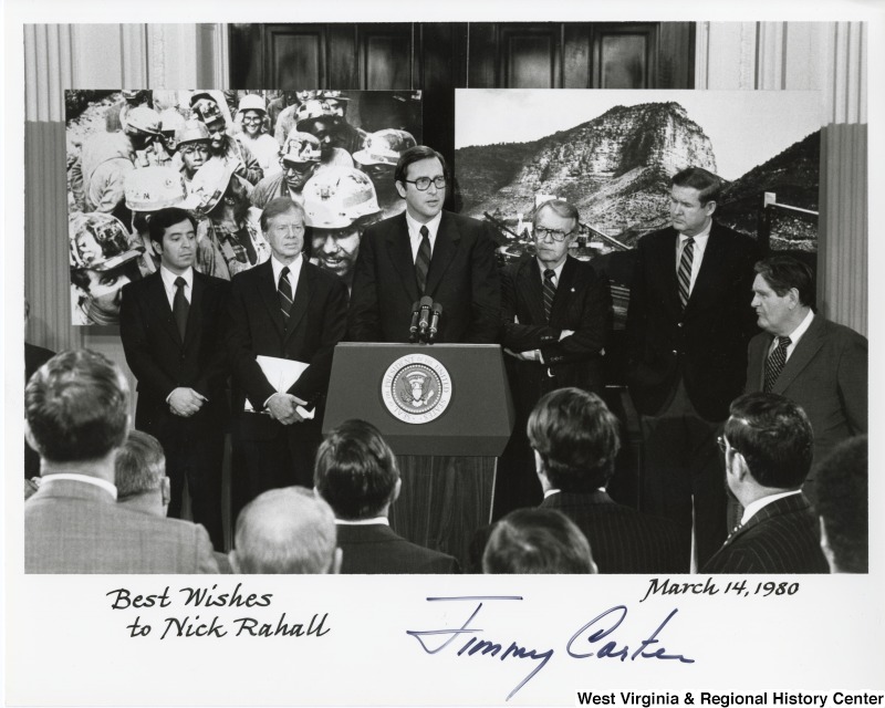 From left to right: Congressman Nick Rahall II (D-WV), President Jimmy Carter, Governor Jay Rockefeller (WV) , Senator Wendell Ford (D-KY), and Congressman John Murtha (D-PA). The photograph was taken for Coal Day at the White House. Governor Jay Rockefeller is speaking to the audience. Behind them are two photographs. One photograph features miners another a mine. The photo signed "Best wishes to Nick Rahall.  Jimmy Carter."