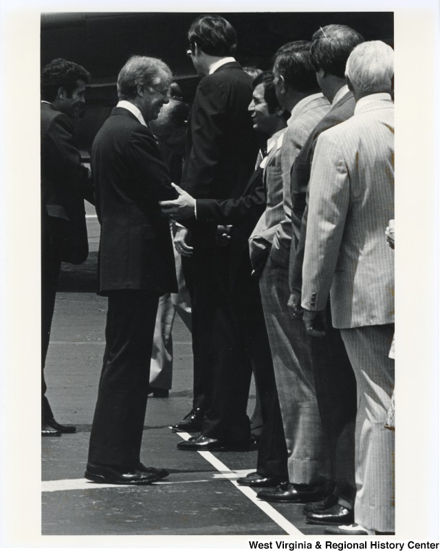 Congressman Nick Rahall II shaking the hand of President Carter. Rahall is in a line with other men. Governor Jay Rockefeller is standing to his right.
