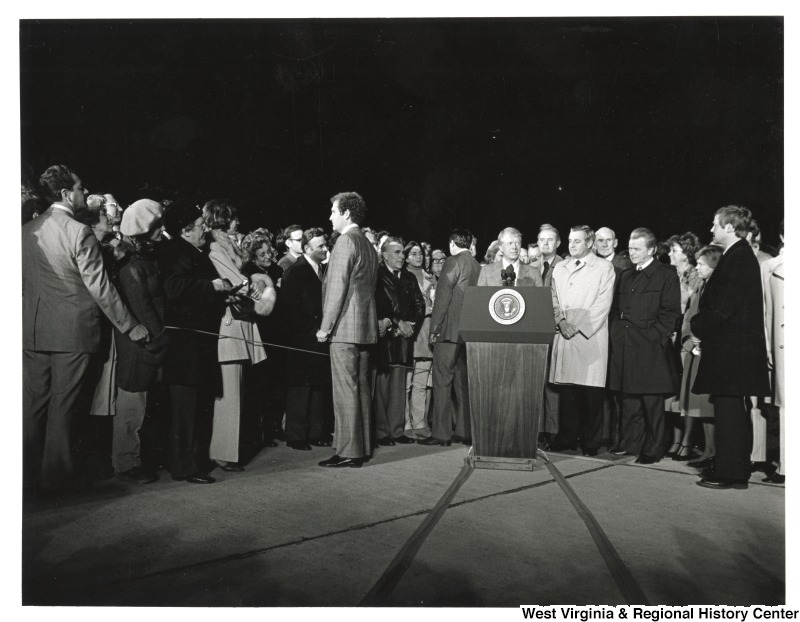 President Carter giving a speaking with a crowd of people behind him. Senator Robert C. Byrd is standing to his left, second person over.