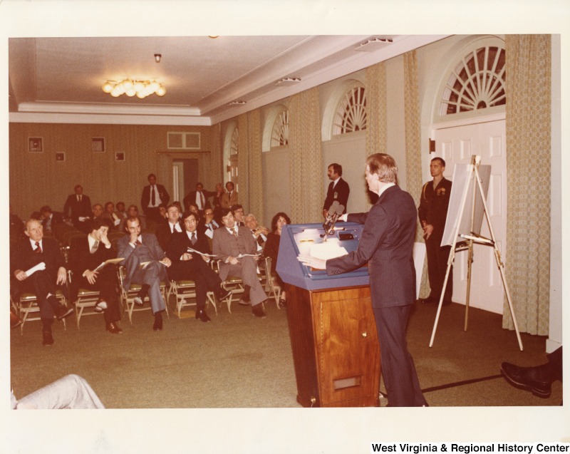 President Jimmy Carter speaking in front of a small group of men. Congressmen Nick Rahall II is seated in the first row, fourth from the left.