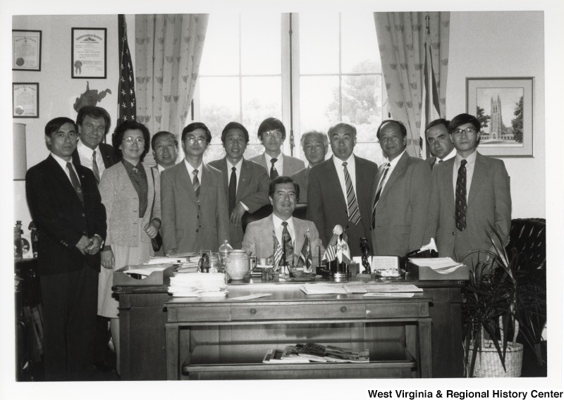 Congressman Nick Rahall II seated at his office desk surrounded by an unidentified group of men and one woman. One of the men is Jack Fairchild.