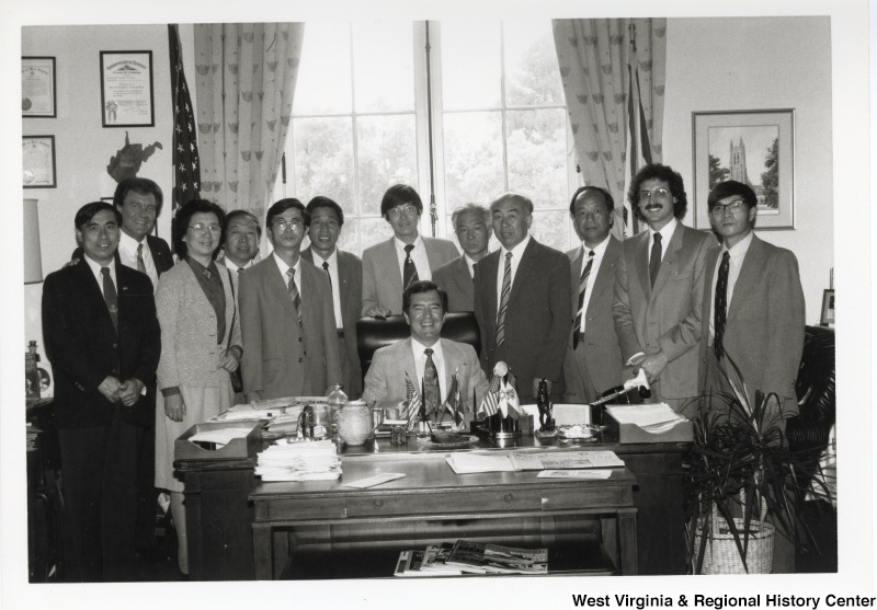 Congressman Nick Rahall II seated at his desk surrounded by an unidentified group of men and one woman. One of the men is Jack Fairchild.