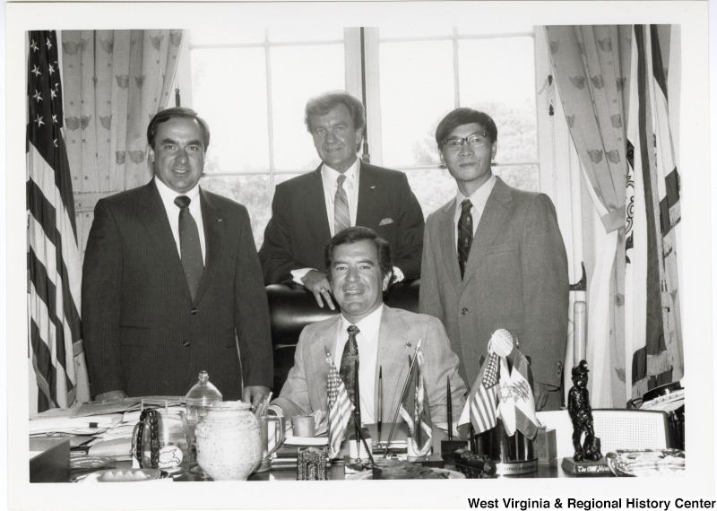 Congressman Nick Rahall II sitting at his desk. Three unidentified men are standing behind him. One of the men is potentially Jack Fairchild.