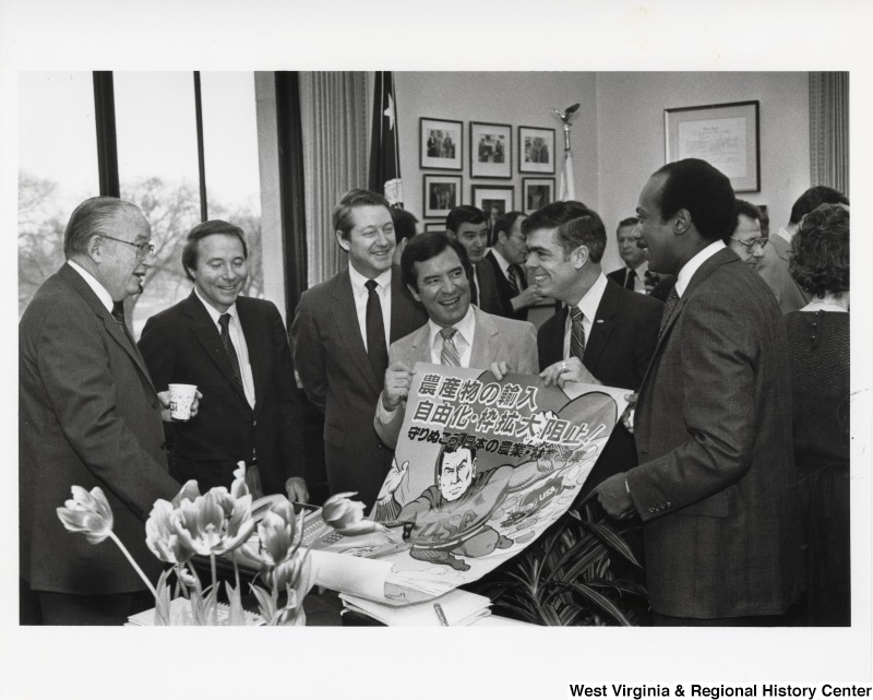 Congressman Nick Rahall II is holding up the left end of a poster with two other men. The poster features a Japanese superhero flying with a USA logo on his chest. One of the men is Jerry Huckabee (D-LA) and another the Secretary of Commerce.