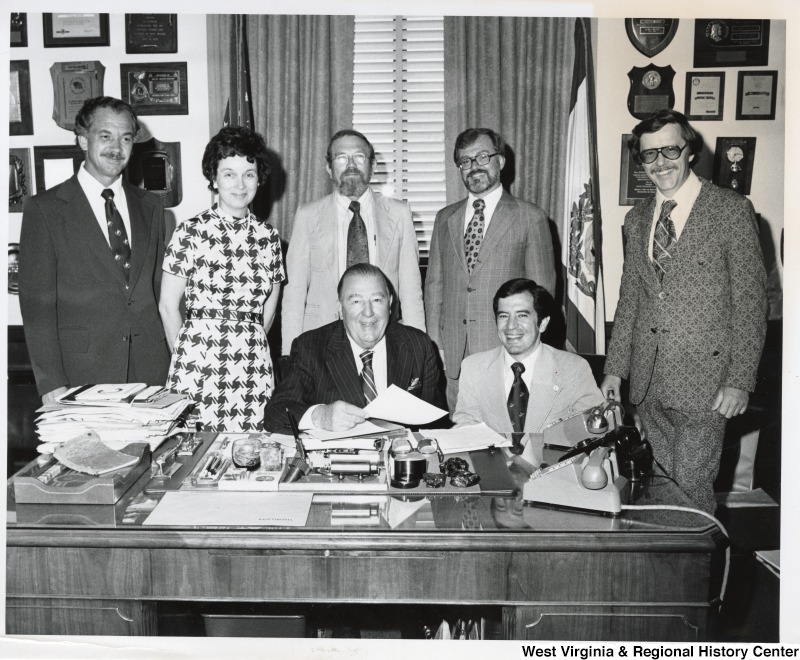 Seated at the desk are Senator Jennings Randolph, and Congressman Nick Rahall II.  Five unidentified people are standing behind them. They are in Senator Jennings Randolph's office.