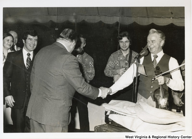 Senator Robert C. Byrd shaking the hand of Senator Jennings Randolph. Byrd is standing in front of a microphone and holding a fiddle. Congressman Nick Rahall II is standing beside Randolph.