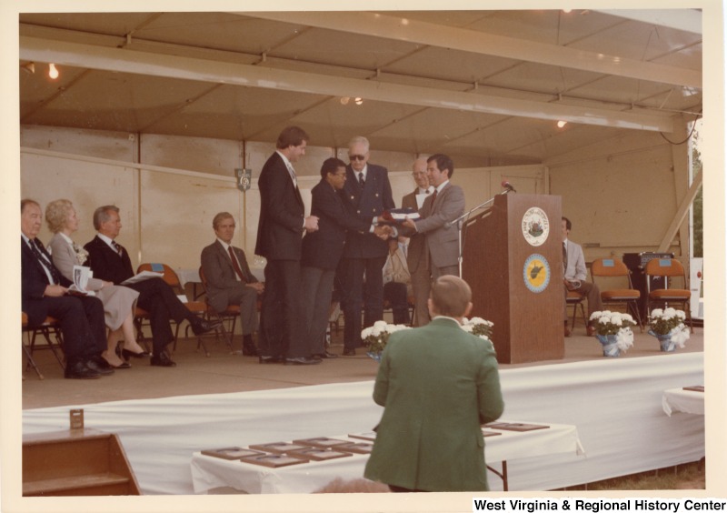 Congressman Nick Rahall II shaking an unidentified mans hand while passing him an unidentified item. Three unidentified people are standing with them on the stage. Senator Jennings Randolph (first on left) is seated with four unidentified men on the stage.