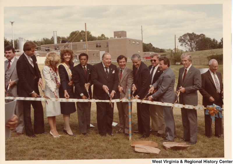 Senator Jennings Randolph (center left) and Congressman Nick Rahall (center right) with ten other unidentified people cutting a ribbon covered in one dollar bills.