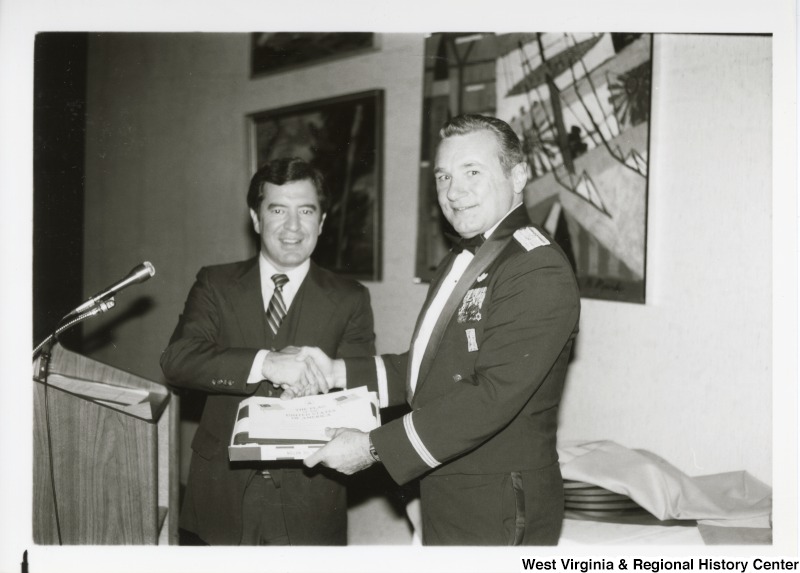 Congressman Nick Rahall II shaking the hand of an unidentified man. They are handing off a box at the same time.