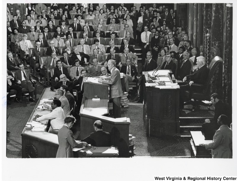 An unidentified man speaking to a auditorium of people. Congressman Nick Rahall II is seated in the first row, second seat from the left.