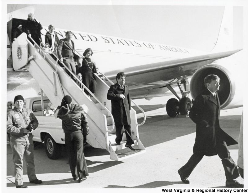 Congressman Nick Rahall II disembarking from a plane. Five other people are descending the stairs behind him. This trip was protentional for the funeral of Congressman Runnels.