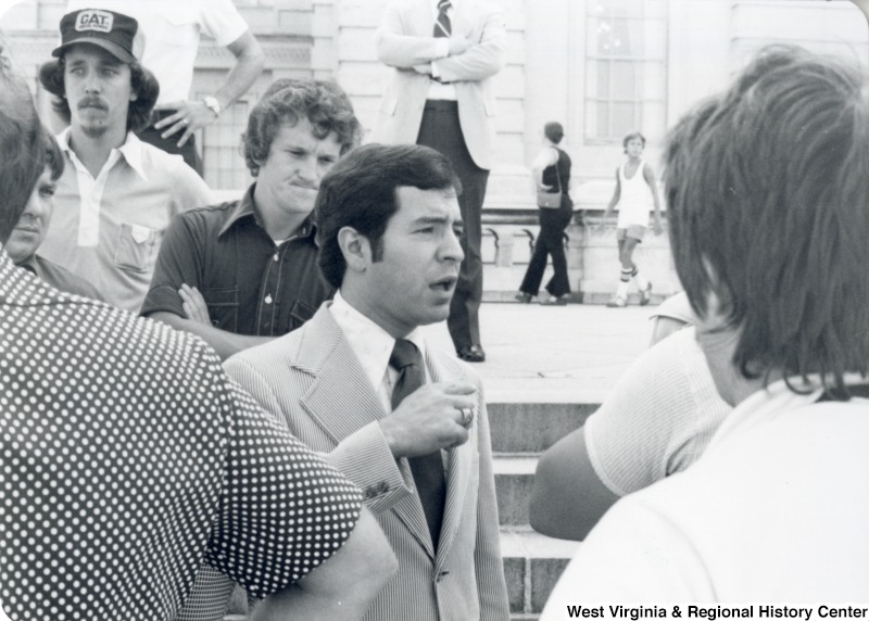 Congressman Nick Rahall II standing in a unidentified group of people. He is speaking to a unidentified person.