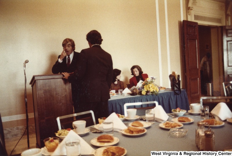 Four unidentified people talking. Two are seated at a table and two are standing by a podium. There is a table with food on it behind them.