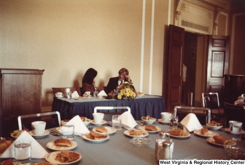 Two unidentified people talking to each other at a table. There is a table with food on it in front of them.