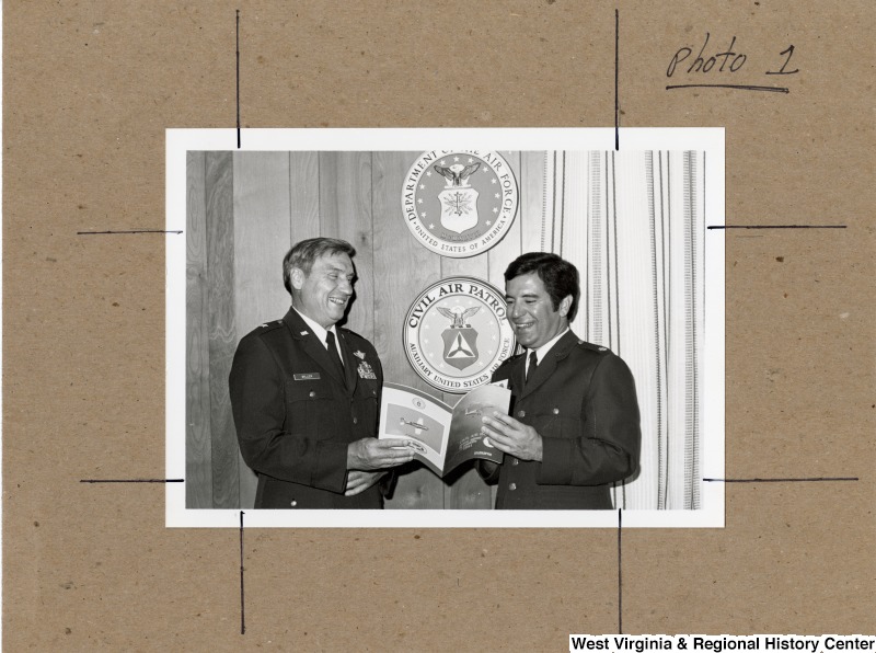 Congressman Nick Rahall II holding a Civil Air Patrol annual report to Congress for 1981 with Brigadier General Miller. Behind them are the insignias for the Department of the AIr Force and the Civil Air Patrol.