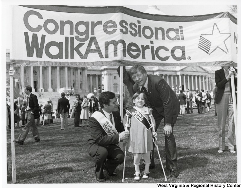 Congressman Nick Rahall II crouched down beside a unidentified little girl with crutches. A unidentified man is standing beside them. They are standing in front of a March of Dimes Congressional WalkAmerica banner.