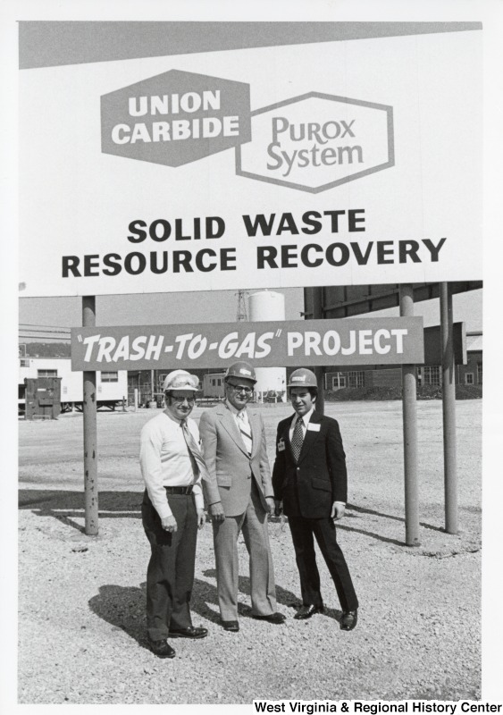 Congressman Nick Rahall II (first on the right) with two unidentified men in front of a Union Carbide and Purox Systems Solid Waste Resource Recovery sign. A smaller sign underneath says 'Trash-to-Gas' project.