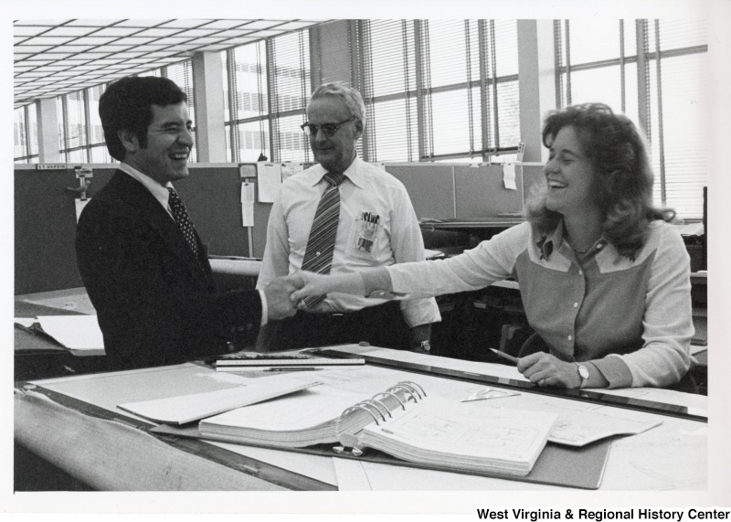 Congressman Nick Rahall II shaking the hand of an unidentified woman who is sitting at a desk. An unidentified man is standing beside Rahall.