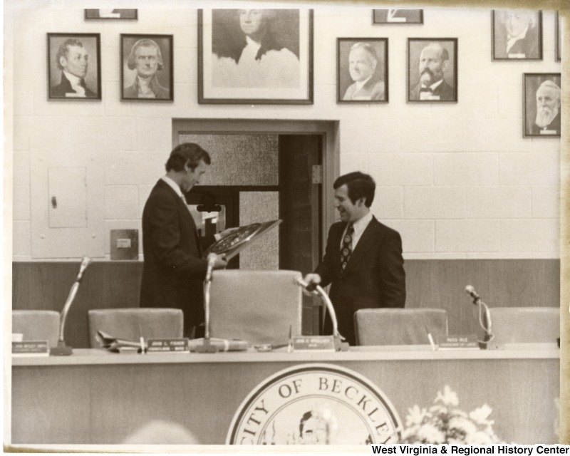 Congressman Nick Rahall II (right) with an unidentified man who is looking at a award. They are standing behind a table with a seal stating City of Beckley.