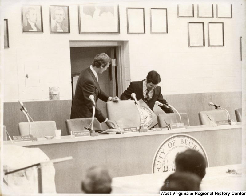 Congressman Nick Rahall II (right) with an unidentified man in front of a unidentified group in Beckley. Congressman Rahall is pulling out a large seal plaque from under the table. There is a table in front of them with a seal stating the City of Beckley.
