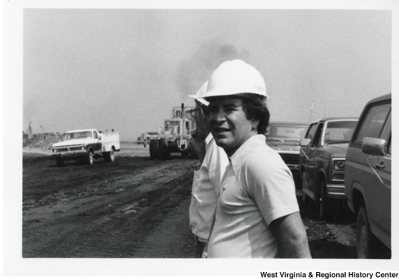 Congressman Nick Rahall II with an unidentified man outside with some machinery. Congressman Rahall is wearing a hard hat.