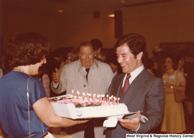 Vicki Rahall presenting a birthday cake to Congressman Nick Rahall II at his 30th birthday fundraising party. Senator Jennings Randolph can be seen in the background between them.