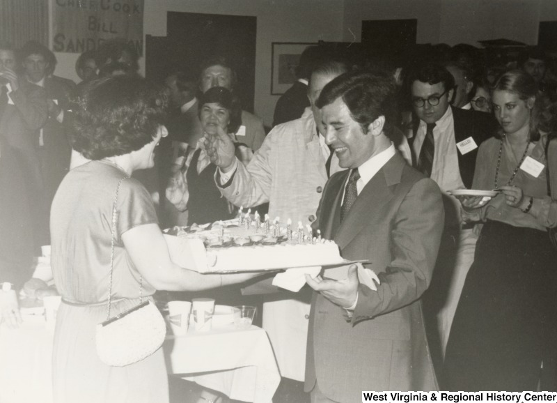 Vicki Rahall presenting a birthday cake to Congressman Nick Rahall for his 30th birthday fundraising party. Senator Jennings Randolph is between them, partially obscured by Rahall.