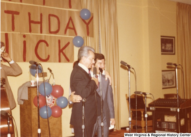 Senator Robert C. Byrd and Congressman Nick Rahall speaking at Congressman Rahall's 30th birthday fundraiser party. A band is standing behind them waiting to play. On the wall is a banner saying "Happy Birthday Nick."