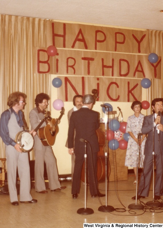 Congressman Nick Rahall II (right, cut off) speaking at his 30th birthday fundraising party. Senator Robert C. Byrd has his back to the crowd speaking with the band. A banner on the wall reads "Happy Birthday Nick".