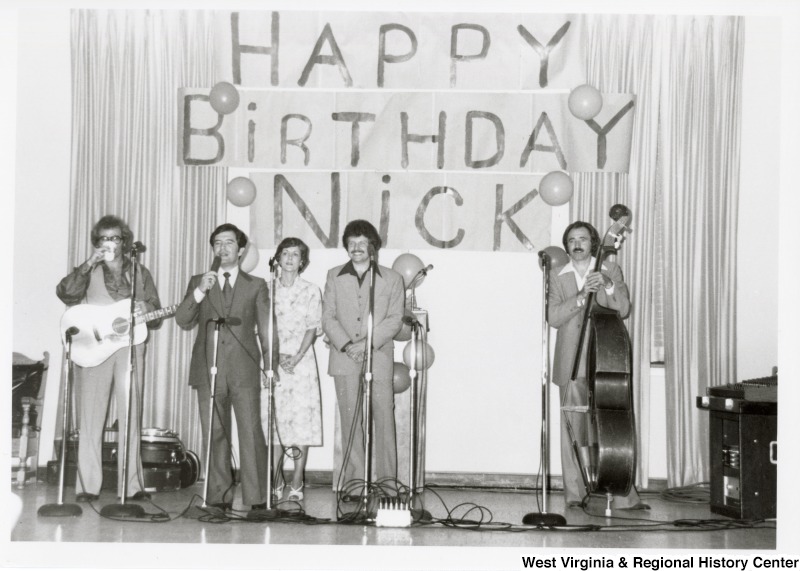 Congressman Nick Rahall II (second from the left) speaking at his 30th birthday fundraising party. An unidentified band is standing with him waiting to play. On the wall is a banner stating "Happy Birthday Nick."
