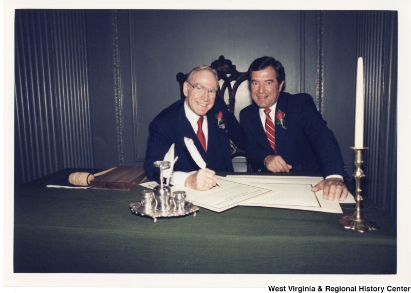 Congressman Jim Wright, Speaker of the House, and Congressman Nick Rahall II sitting at a desk. Congressman Wright is signing a document with a feather quill.
