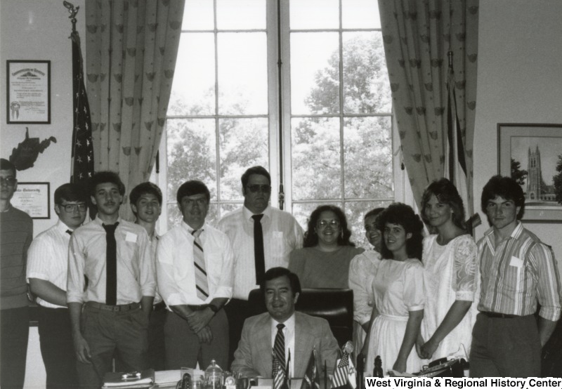 Congressman Nick Rahall II seated at his desk surrounded by an unidentified group of men and women.