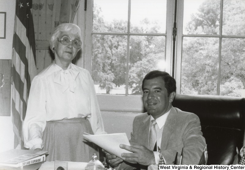 Congressman Nick Rahall II with an unidentified woman in his office. Rahall is seated at his desk and is holding some paper.