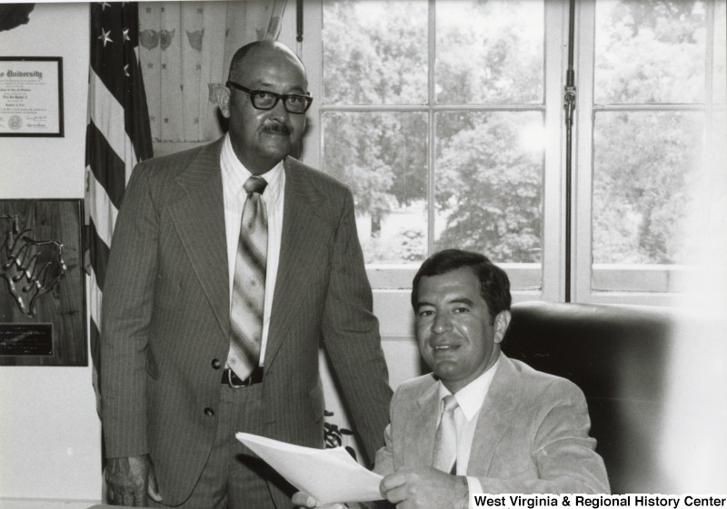 Congressman Nick Rahall II in his office with an unidentified man. Rahall is seated at his desk and holding a piece of paper.