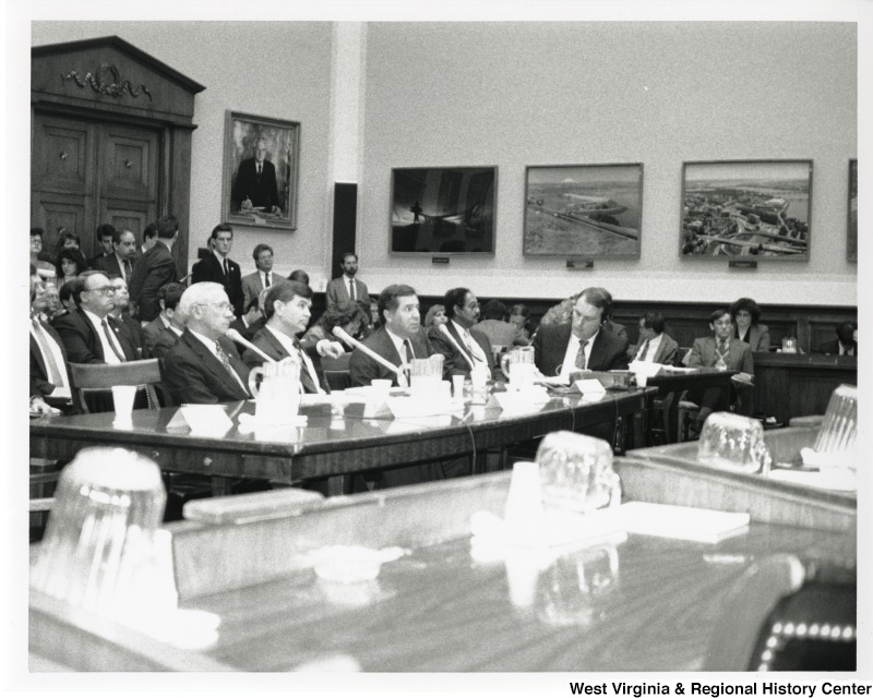 Congressman Nick Rahall II (seated third from the left) in an unidentified meeting or hearing. Four unidentified men are sitting with Rahall. Behind them is a group of people sitting and standing listening to the meeting.