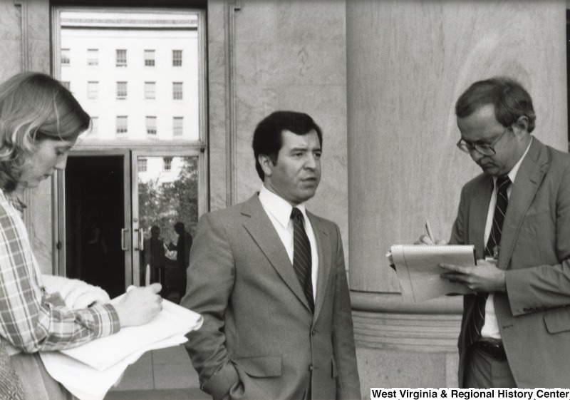 Congressman Nick Rahall II (center) speaking to an unidentified man and woman who are taking notes.
