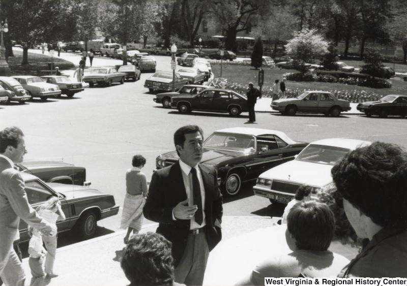 Congressman Nick Rahall II outside speaking to an unidentified group of people.  Cars can be parked along the street behind Rahall.