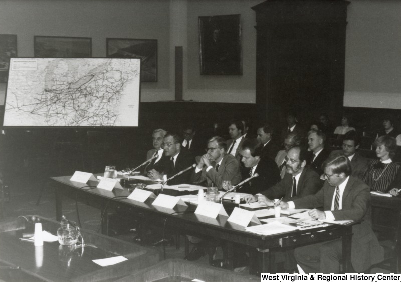 An unidentified man speaks during a committee meeting or hearing. Five unidentified men sit at the table with him.